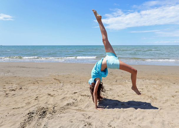 young beautiful girl performs a wheel with her legs in the air by the sea on the sandy beach in summer - Foto, Imagem