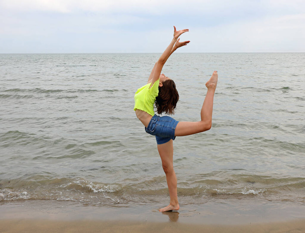 joven esbelta adolescente realiza un ejercicio de gimnasia artística con la pierna detrás de la espalda llamada ECART en la orilla del mar - Foto, imagen