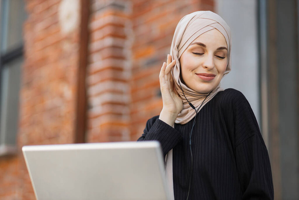 Close up portrait of arabic muslim young women wearing hijab listening to music with headset, having coffee break and working on laptop outdoor on city background. - Valokuva, kuva