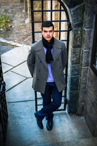 Waiting for You. Young man wearing gray coat, black scarf, blue jeans, black leather shoe, standing by steel gate in New York City in winter, looking up, waiting - Foto, Bild