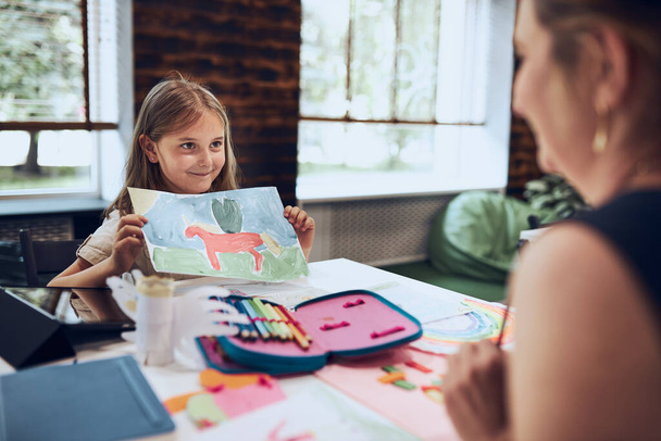 Girl presenting her artwork teacher. Woman assisting schoolgirl during classes at primary school. Child drawing picture sitting at desk in classroom. Girl drawing pictures. Learning at primary school. Elementary education. Back to school - Фото, изображение