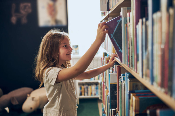 Schoolgirl choosing book in school library. Smart girl selecting literature for reading. Books on shelves in bookstore. Learning from books. School education. Benefits of everyday reading. Child curiosity. Back to school - Φωτογραφία, εικόνα