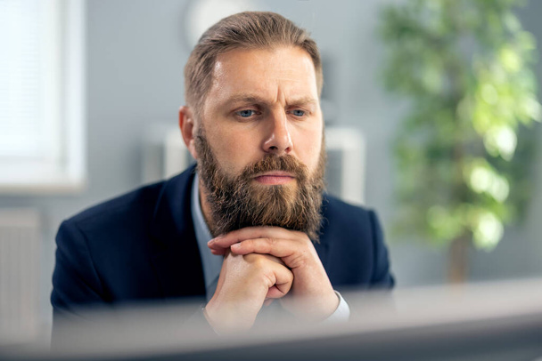 Thoughtful caucasian man with trendy beard and haircut looking on pc screen while working at bright modern office. New ideas, dedication to work and business concept. - Foto, Imagem