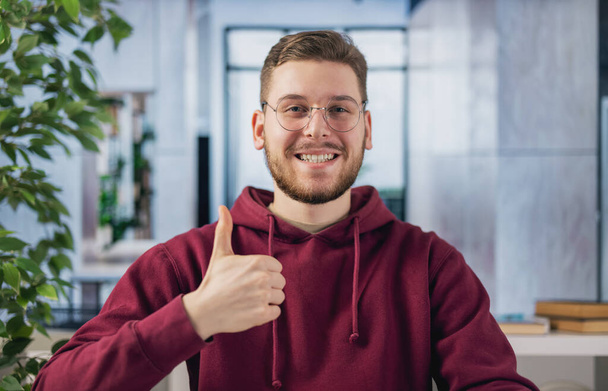 Portrait of smiling caucasian man in glasses and hoodie gesturing thumb up while sitting at workplace. Cheerful expression and winner gesture. - 写真・画像