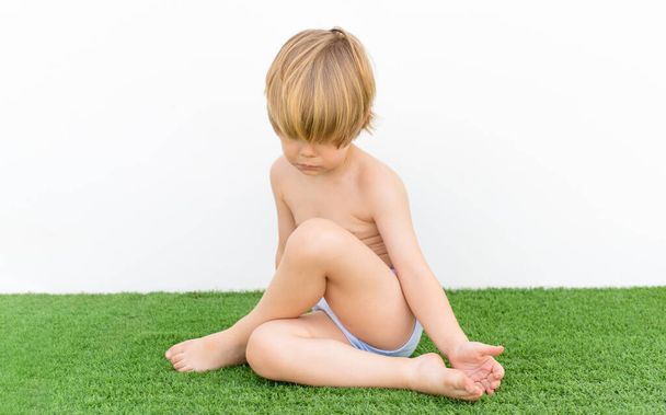 Cute little boy in swimsuit sits on a grass in a summer garden. - Фото, изображение