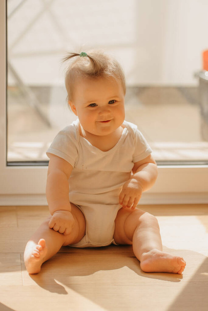 Uma menina gentil de 7 meses está sentada perto de uma porta da varanda em um bodysuit em casa. Um bebê bonito está sorrindo à luz do sol. - Foto, Imagem