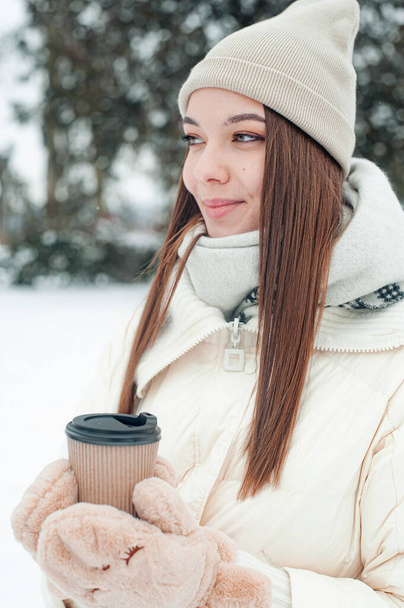  Πορτρέτο της γυναίκας πίνοντας τσάι σε εξωτερικούς χώρους. Νεαρή γυναίκα με ένα φλιτζάνι ζεστό ποτό διασκεδάζει στο πάρκο σε χιονισμένο καιρό. - Φωτογραφία, εικόνα