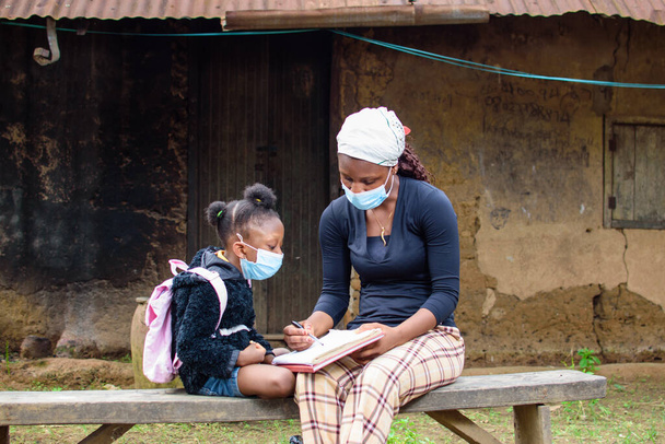 Een Afrikaanse moeder of leraar buiten een dorp modderhuis, het helpen van een meisje kind te repareren haar neus masker tijdens persoonlijke studie voor uitmuntendheid in haar onderwijs - Foto, afbeelding