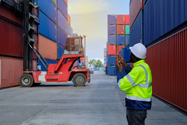 African American male worker in safety uniform and hardhat use walkie-talkie, work at logistics terminal with many stacks of containers, loading control shipping goods, cargo transportation industry. - Photo, Image