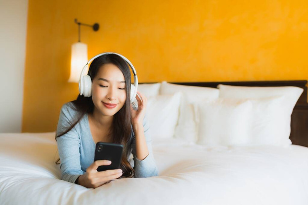 Πορτρέτο όμορφη νεαρή Ασιάτισσα γυναίκα χρησιμοποιώντας κινητό τηλέφωνο με ακουστικά για να ακούσετε μουσική στο εσωτερικό του υπνοδωματίου - Φωτογραφία, εικόνα