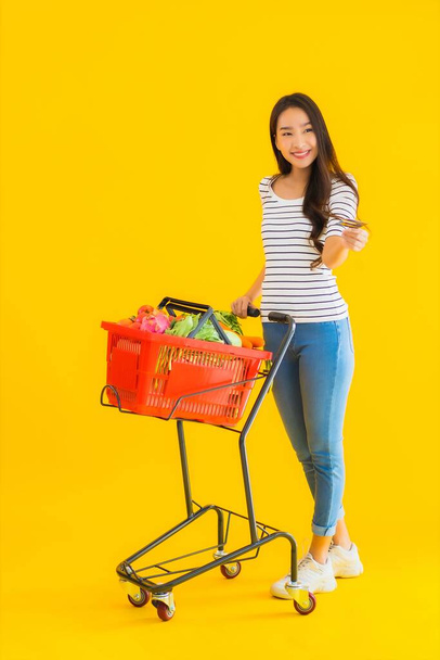 Porträt schöne junge asiatische Frau mit Einkaufswagen aus Supermarkt in Einkaufszentrum auf gelbem isolierten Hintergrund - Foto, Bild