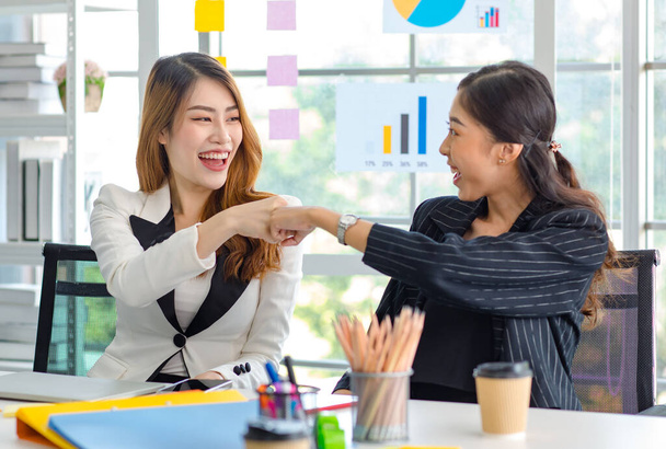 Two happy cheerful Asian millennial professional successful female businesswomen in formal suit sitting together holding hands fists bumping celebrating job agreement deal done at office workstation. - Foto, Bild