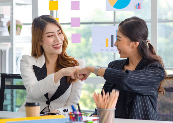 Two happy cheerful Asian millennial professional successful female businesswomen in formal suit sitting together holding hands fists bumping celebrating job agreement deal done at office workstation. - Foto, Imagem