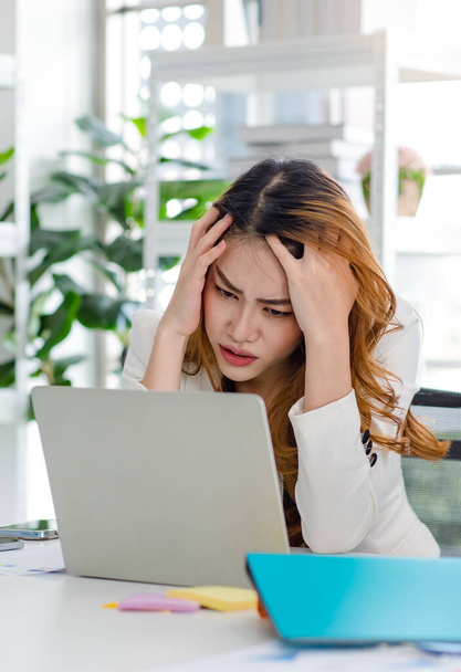 Szoros felvétel ideges stresszes depressziós aggódó boldogtalan ázsiai millenniumi ráncoló arc üzletasszony alkalmazott ülni kéz a kézben a fejét, miközben dolgozik laptop notebook számítógép vállalati irodában. - Fotó, kép