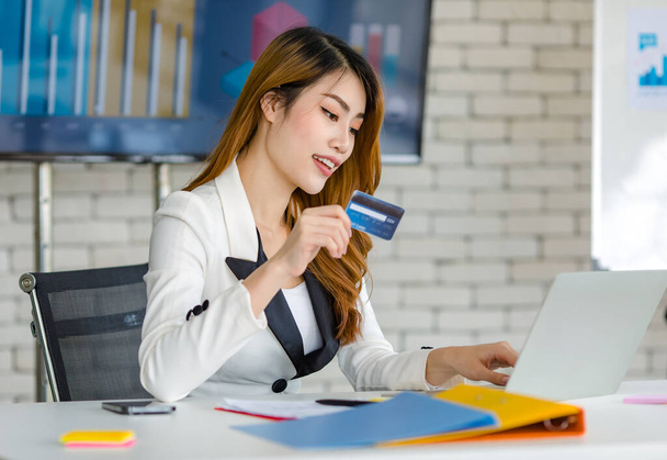 Asian young happy cheerful beautiful millennial professional successful female businesswoman in formal suit sitting smiling holding credit card in hand shopping online via laptop notebook computer. - Foto, imagen