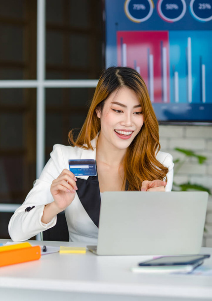 Asian young happy cheerful beautiful millennial professional successful female businesswoman in formal suit sitting smiling holding credit card in hand shopping online via laptop notebook computer. - Photo, Image
