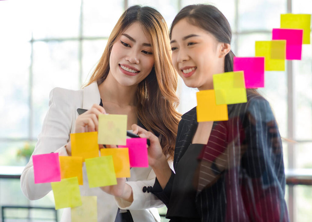 Asian young happy cheerful millennial professional successful businesswoman employee in formal suit standing smiling discussing with colleague writing post it paper on glass board in meeting room. - 写真・画像