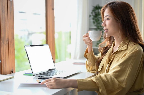 Mooie Aziatische zakenvrouw of vrouwelijke manager ontspant nippen een ochtend koffie tijdens het werken aan haar bureau in het kantoor. - Foto, afbeelding