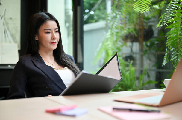 Mujer de negocios asiática segura y exitosa o gerente ejecutiva en traje formal mirando y revisando un informe de negocios en la carpeta en su escritorio de oficina. - Foto, Imagen