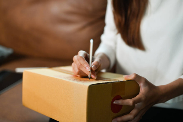 Μια γυναίκα γράφει μια διεύθυνση στο χαρτόκουτο. Μια γυναίκα ιδιοκτήτρια ηλεκτρονικού καταστήματος ετοιμάζει ένα πακέτο αποστολής. περικοπή και εστίαση εικόνας του χεριού. - Φωτογραφία, εικόνα