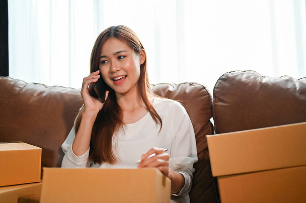 Beautiful millennial asian woman sitting on sofa with cardboard boxes and talking on the phone with house moving service. House moving, relocation concept. - Photo, Image