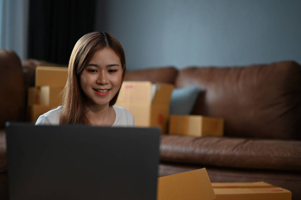 Schöne Millennial asiatische KMU-Geschäftsinhaberin mit Laptop-Computer bei der Vorbereitung eines Kunden Aufträge im Wohnzimmer. - Foto, Bild