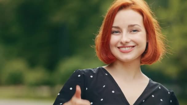 Stylish young pretty girl attractive caucasian redhead teen woman girlfriend cheerful smiling friendly looks at camera puts thumb up gesture recommends best choice shows everything fine stands outside - Filmmaterial, Video