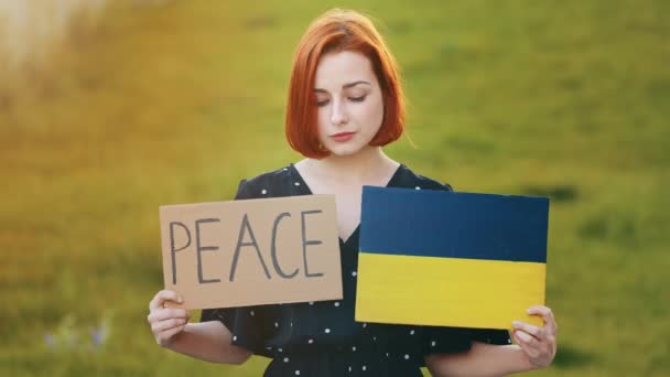 Sad redhead woman young patriotic girl activist stand outdoors hold two banners with lettering word peace cardboard drawing in blue yellow ukrainian flag colours asking help for Ukraine anti war sign - Video, Çekim