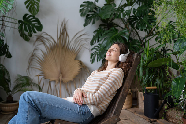 Beautiful young woman in wireless headphones sits with close eyes on wooden chair in urban jungle garden relax space. Calm female enjoying good meditative music, online radio, audiobook or podcasts. - Photo, image