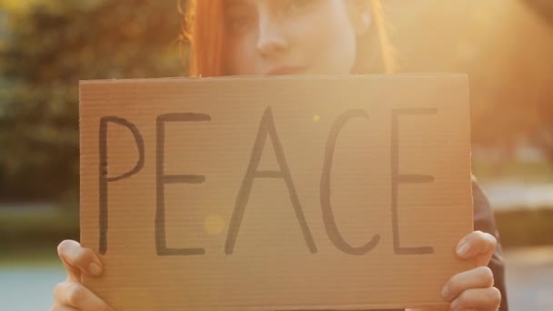 Close up female hands hold cardboard with written word peace symbolic banner attractive young woman pretty redhead girl stand in park demonstrate protesting slogan peaceful poster anti victim placard - Imágenes, Vídeo