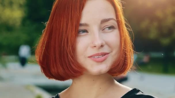 Közelkép emberi nő arc vörös hajú nő barátnő természetes smink tökéletes tiszta bőr mosolygós fogas mosoly gyönyörű vonzó modell lány hölgy flörtölés pózol a városi parkban napsütés háttér - Felvétel, videó