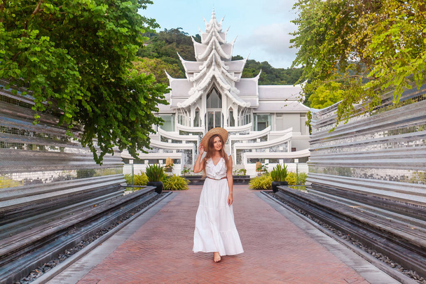 Traveler woman in white dress walking in front of a beautiful white temple in thai style. Travel and vacation in Asia. Young woman in hat near beautiful buddhist temple on Phuket Island in Thailand - Φωτογραφία, εικόνα
