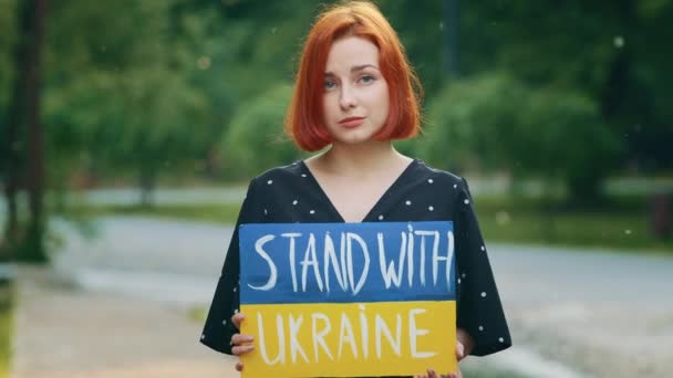 Sad redhead girl hold cardboard with written inscription phrase stand with Ukraine shows patriotic banner painted in ukrainian national blue yellow colors upset anxious young woman ask active support - Footage, Video
