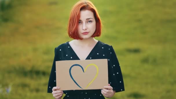 Unhappy sad redhead woman hold artwork colorful cardboard drawing blue yellow love shape heart romantic symbol demonstrate tenderness feelings attractive young pretty girl stands outdoors in sunlight - Footage, Video