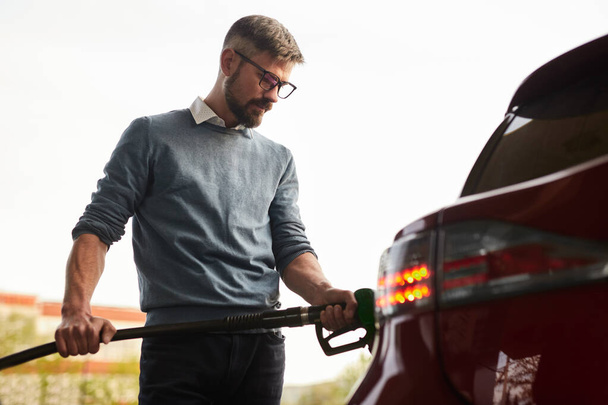 Low angle of concentrated adult bearded male client in smart casual clothes and eyeglasses refueling modern car using gas pump nozzle at filling station - 写真・画像