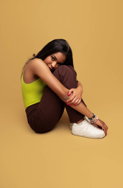 Side view of shy teen model in trendy clothes with black hair embracing knees and looking and camera while sitting against ocher background - Фото, изображение