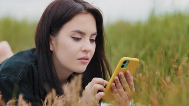 Кавказька вродлива жінка, що лежить на траві, насолоджується вільним часом на самоті, використовуючи смартфон-прилад для розмов з повідомленням в Інтернеті сторінки в соціальних мережах, дивлячись на смішні зображення, які читають новини. - Кадри, відео