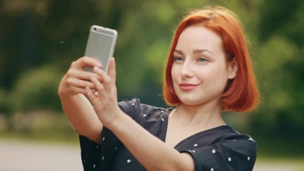 Portrait stylish beautiful girl caucasian attractive redhead woman blogger female model stands outside in park holds smartphone takes selfie photo on mobile camera records vlog filming video for blog - Footage, Video