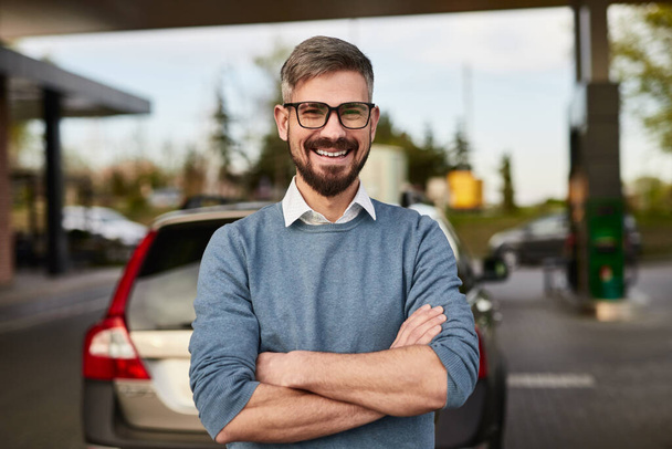 Cheerful adult male customer with beard in smart casual outfit and eyeglasses smiling happily, and looking at camera while standing near mode SUV car at gas station with crossed arms - Фото, зображення