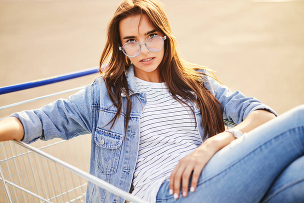 Self assured young female millennial with long brown hair in casual outfit and trendy, eyeglasses sitting in shopping cart and looking at camera on street on sunny day - Fotografie, Obrázek