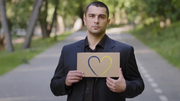 Sad serious adult caucasian man unhappy guy businessman worker stand outside hold colorful painted cardboard drawing love shape heart in blue yellow national colors Ukraine symbol of romantic feelings - Video, Çekim