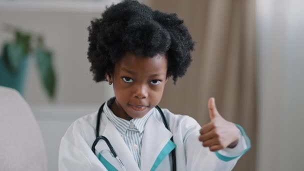 Close-up little cute kid girl in medical gown looking at camera posing indoors smiling pretending be doctor plays nurse showing thumb up gesture approval promotes dream job future profession concept - Záběry, video