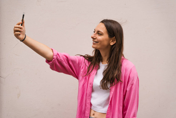 Portrait of a caucasian brunette smiling woman in casual pink shirt taking selfie, look at smartphone front camera, photographing, record video message post online. Isolated on a white background. - Fotó, kép