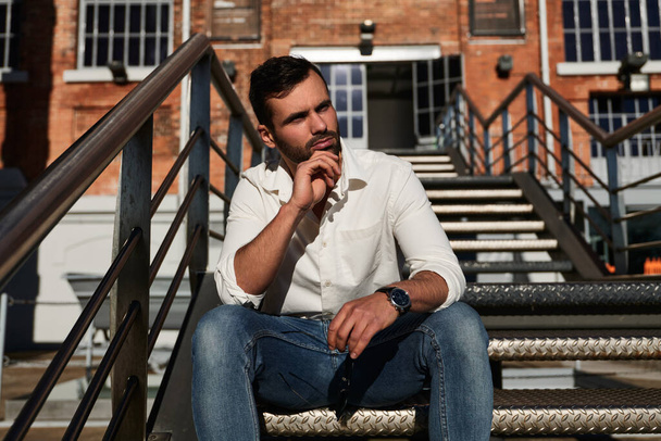 Pensive ethnic male in white shirt and jeans touching chin and looking away while sitting on metal steps on city street in daytime - 写真・画像