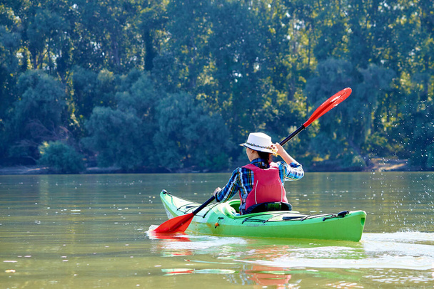ドナウ川で緑のカヤックを漕ぐエレガントなわら帽子のカンチプライヤーの女性。夏のウォータースポーツ - 写真・画像