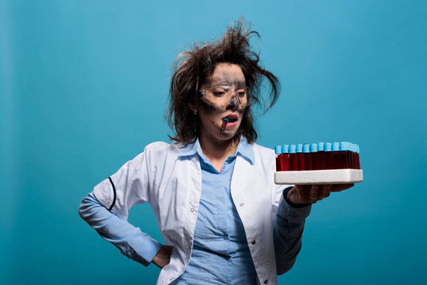 Mad biochemistry specialist with dirty face and messy hairstyle holding tray with glass test tubes while on blue background. Crazy scientist with test tube between teeth looking careless at glassware. - Valokuva, kuva