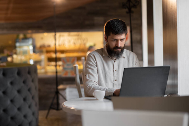 Kafede çalışan genç sakallı bir adam dizüstü bilgisayar kullanıyor, kafede çalışan serbest çalışan bir işçi, kafenin içinde çalışan bir adam. - Fotoğraf, Görsel