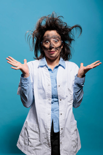 Insane and silly looking crazy scientist with messy hairstyle and dirty face does not understand where experiment went wrong. Mad chemist with goofy expression standing on blue background. Studio shot - Foto, Bild