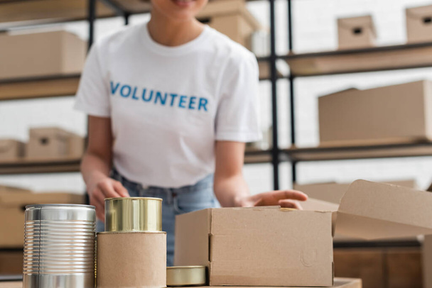 partial view of blurred african american woman working near canned foodstuff in volunteer center - Photo, Image