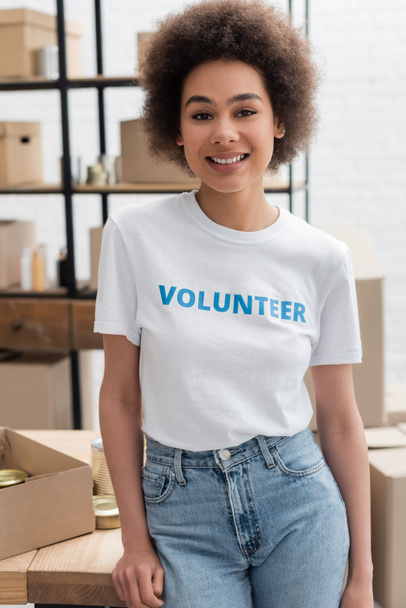 happy african american woman in t-shirt with volunteer lettering looking at camera in charity center - Foto, imagen